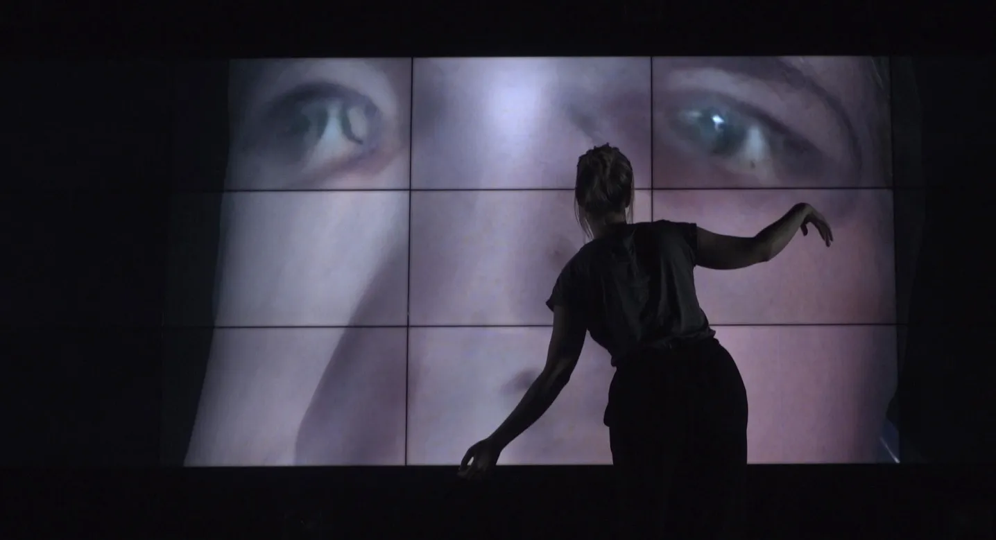 Work-in-progress preview of SELF ABSORBED by Tim Murray-Browne. A woman stands in front of a large video wall striking a pose with her arms. On the video wall is a 3D render of a distorted male face.