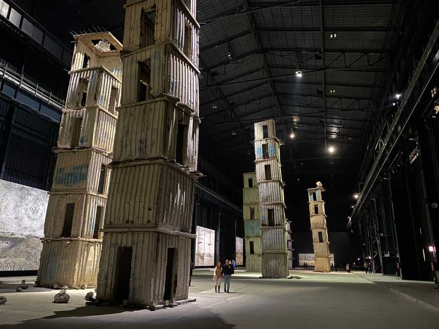 A photo of Anselm Kiefer's installation "The Seven Heavenly Palaces" at Hangar Bicocca. It shows concrete towers apparently precariously stacked up to many stories high, with two people standing next to them demonstrating the scale.