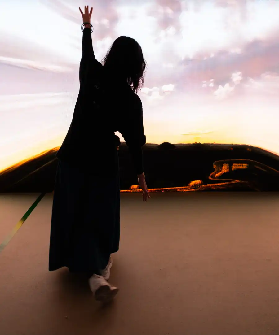 The installation Self Absorbed by Tim Murray-Browne. A woman stands tilted with one arm in the air in front of a wall-sized LED screen showing clouds and a landscape. Photo: Camila Picolo